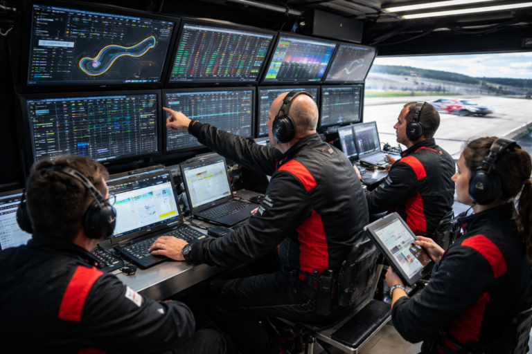 Motorsports team members seated at a pit wall workstation reviewing telemetry, timing screens, and performance data while cars run on track.